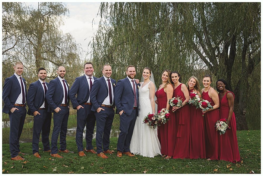 Wedding party poses outdoors. Groomsmen in navy suits, bridesmaids in red dresses, bride in white dress, holding bouquets.