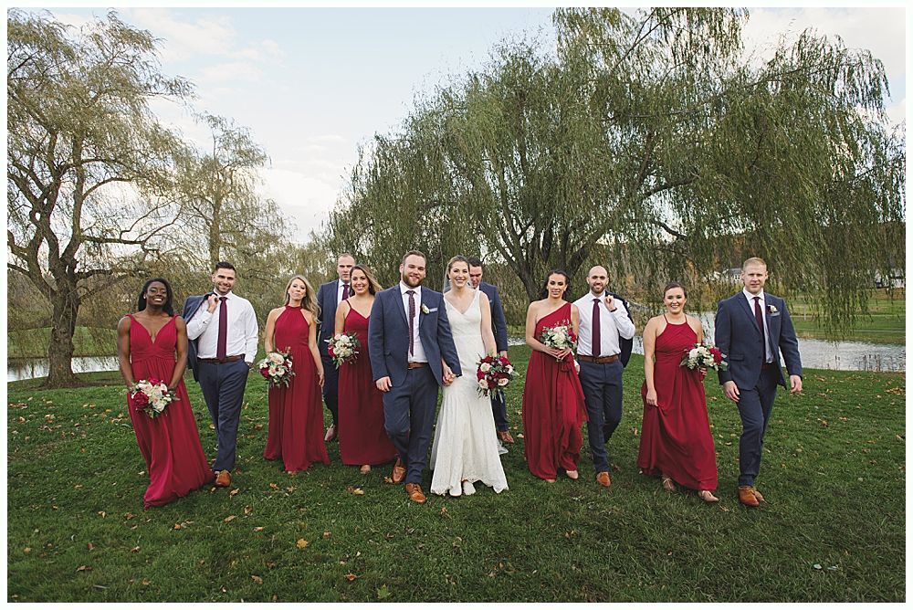 Wedding party walking on a grassy lawn; bride and groom in center, bridesmaids in red, groomsmen in navy.