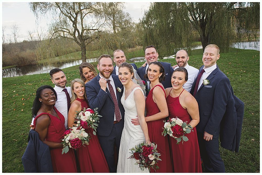 Wedding party smiling outdoors near water. Bride and bridesmaids in red, groom and groomsmen in navy.