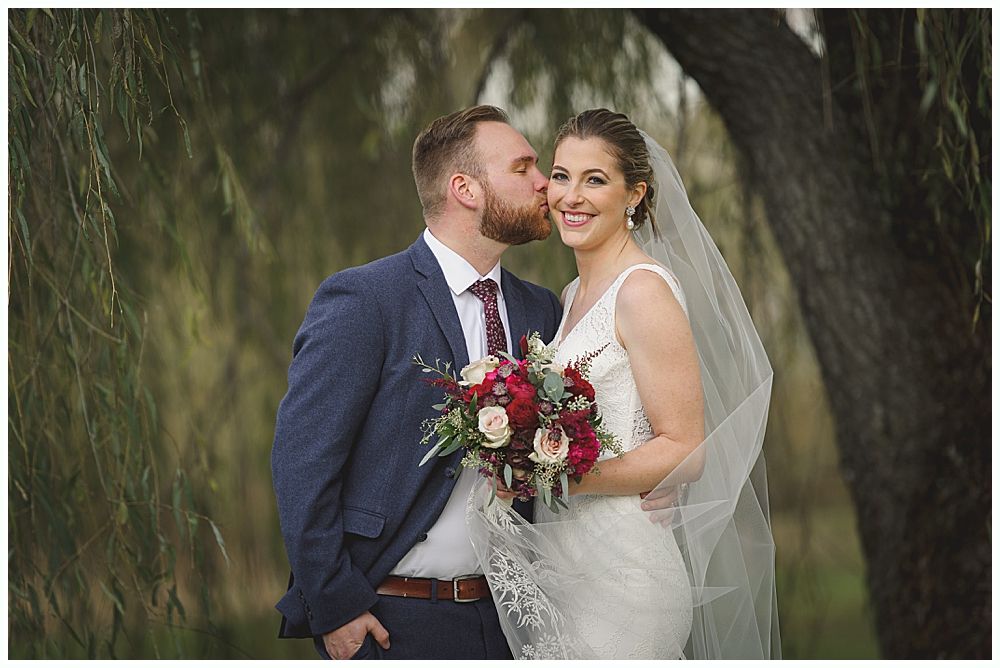 Groom kissing bride's cheek; she holds flowers. They are outdoors, near a tree. She wears a veil and dress, he wears a suit.