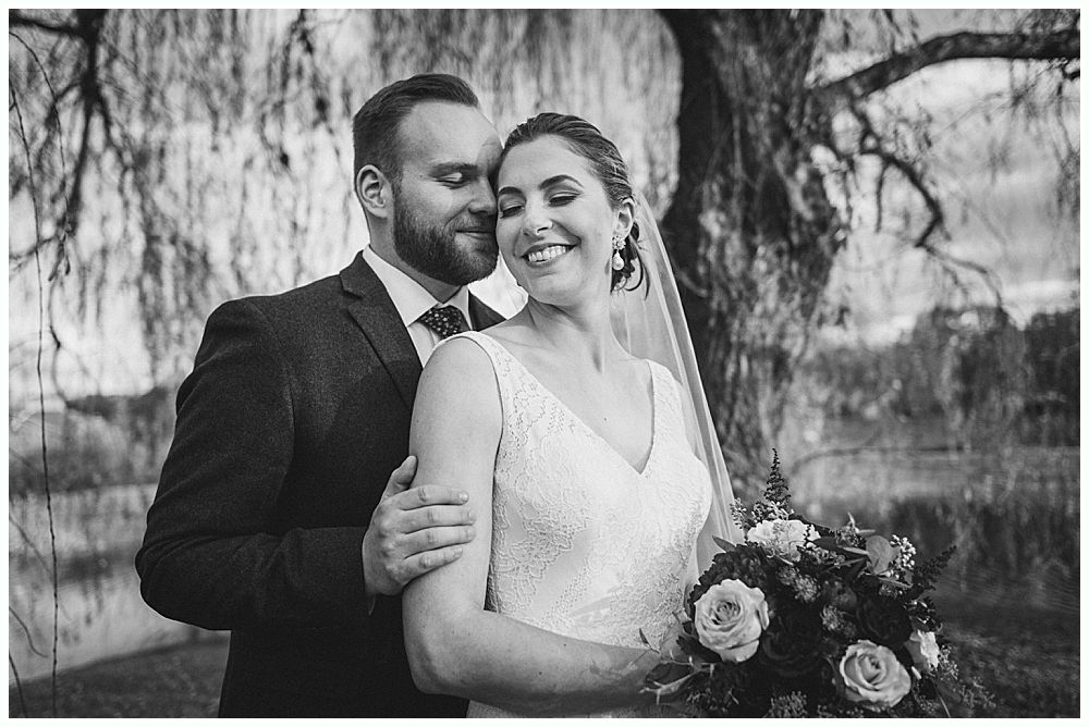 Bride and groom embracing, smiling, holding flowers. Trees and water in the background. Black and white.
