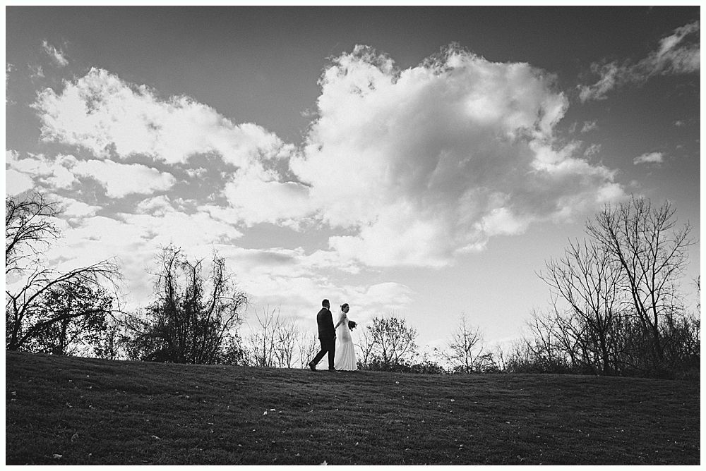 Bride and groom walk hand-in-hand across a grassy hilltop; black and white photograph with a cloudy sky backdrop.