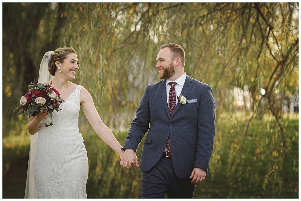 Bride and groom holding hands, smiling. She wears a white dress and veil, he wears a blue suit. Outdoors, near trees.