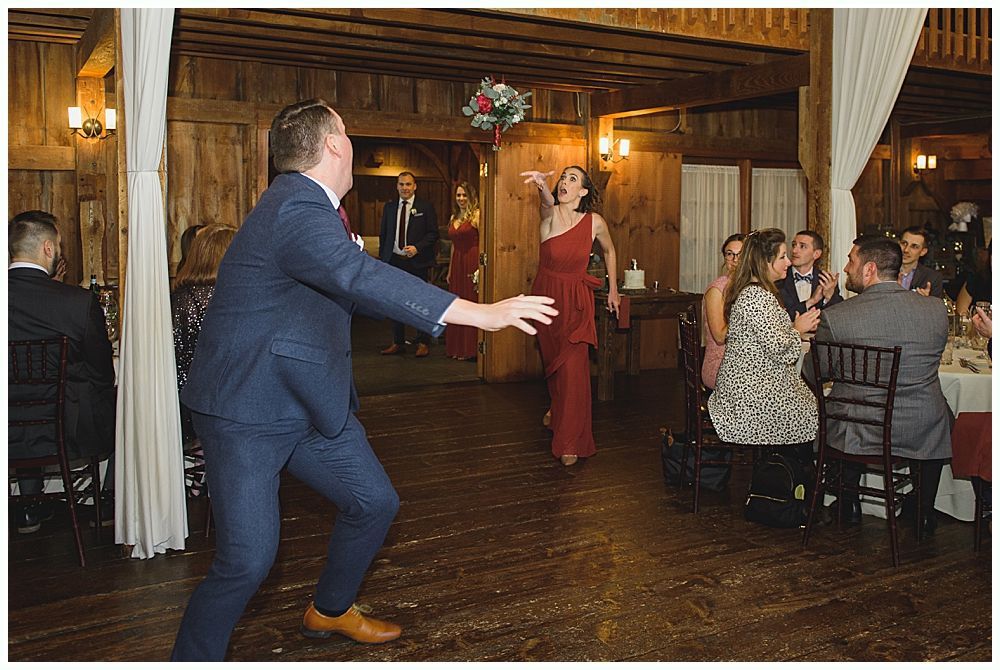 Woman in red gown throws bouquet at wedding reception. Man in suit reaches for it. Wooden interior.