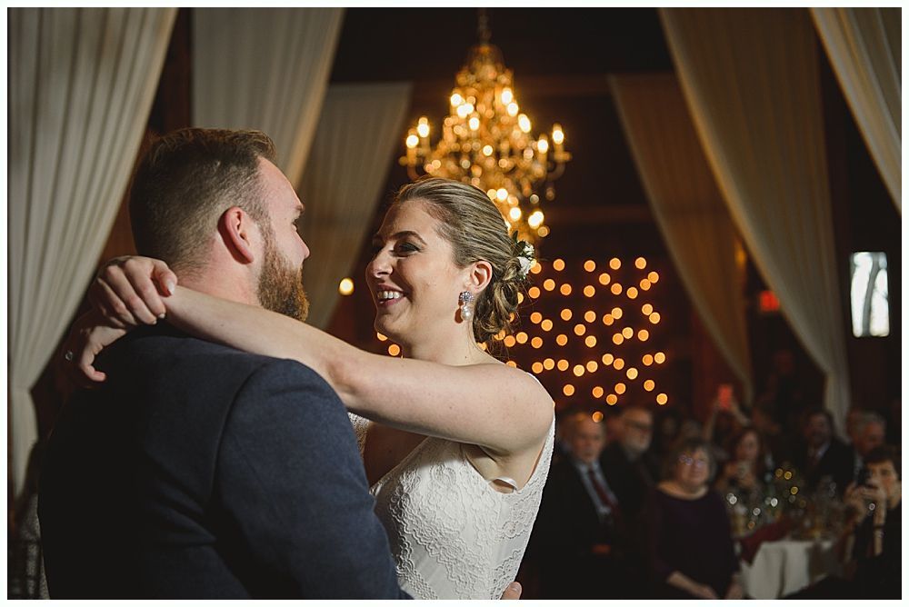 Couple dances at a wedding reception; chandelier and string lights in background.