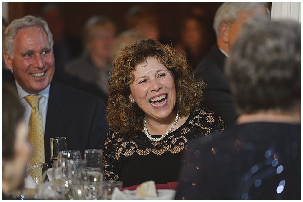Woman laughs at a formal dinner, seated next to a smiling man with a yellow tie; table setting.