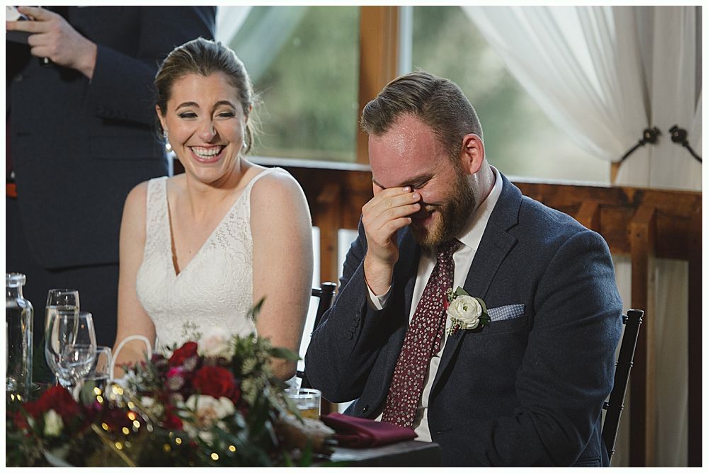 Wedding couple laughing during a reception. The groom covers his face, and the bride smiles widely.
