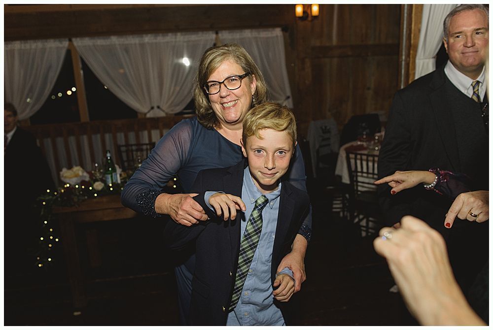 Woman hugs boy wearing a tie and blazer in a warmly lit room. Both smile.