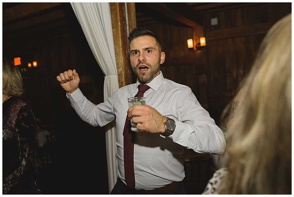 Man dancing with a drink, wearing a dress shirt and tie, inside a warmly lit venue.