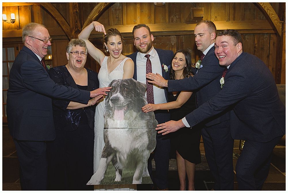 People at a wedding posing with a large cardboard cutout of a dog. Interior setting.