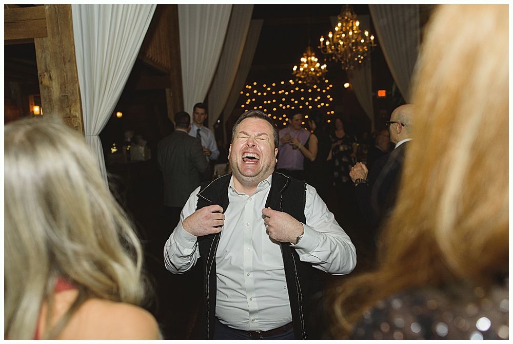Man laughing, dancing at party, wearing a black vest and white shirt. Other people and chandeliers in background.