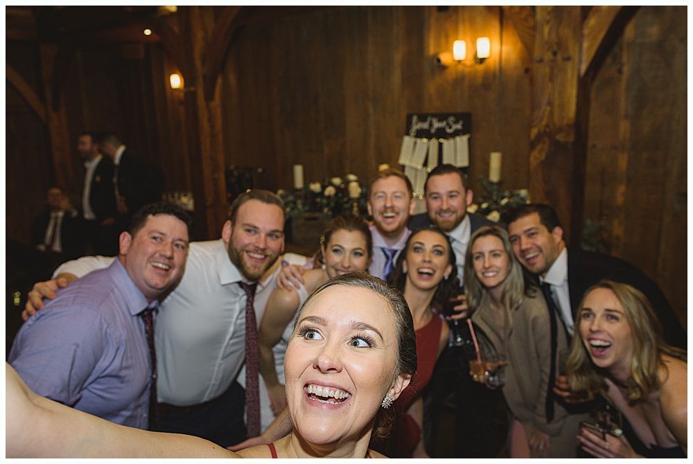 Group of people smiling, taking a selfie inside a rustic wooden barn at a wedding reception.