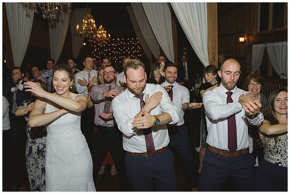 Wedding reception: Bride and guests dance, arms crossed. Wooden interior, festive lights.