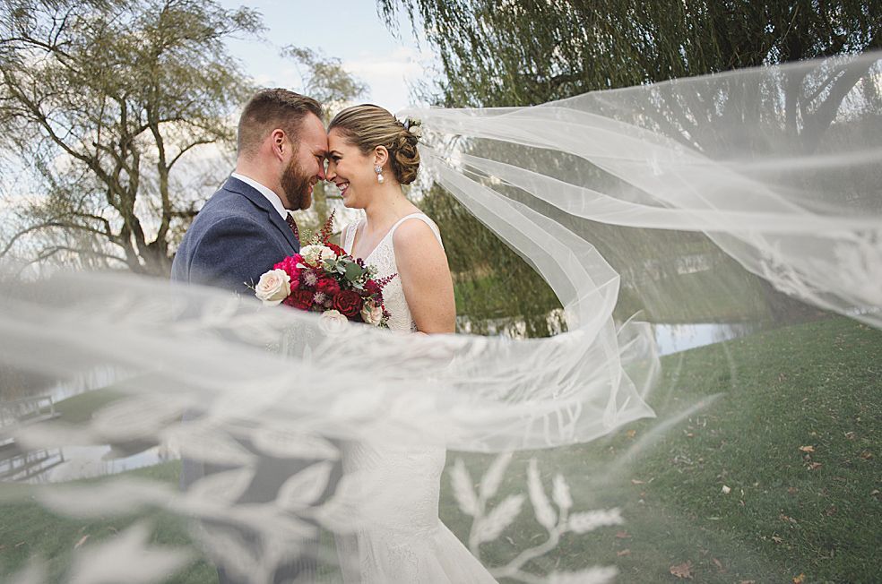 Bride and groom embrace, veil billowing. They are outdoors, holding flowers, smiling.