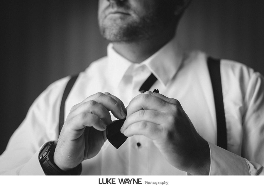Man in formal wear adjusting a bow tie; suspenders visible, black and white photo.