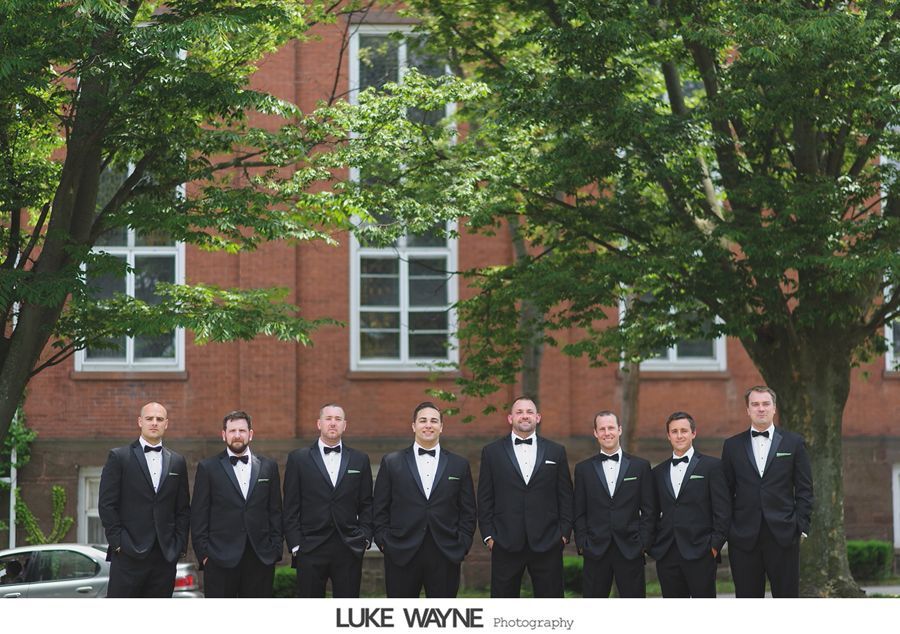 Groomsmen in black tuxedos stand in front of a brick building and trees, posing for a photo.