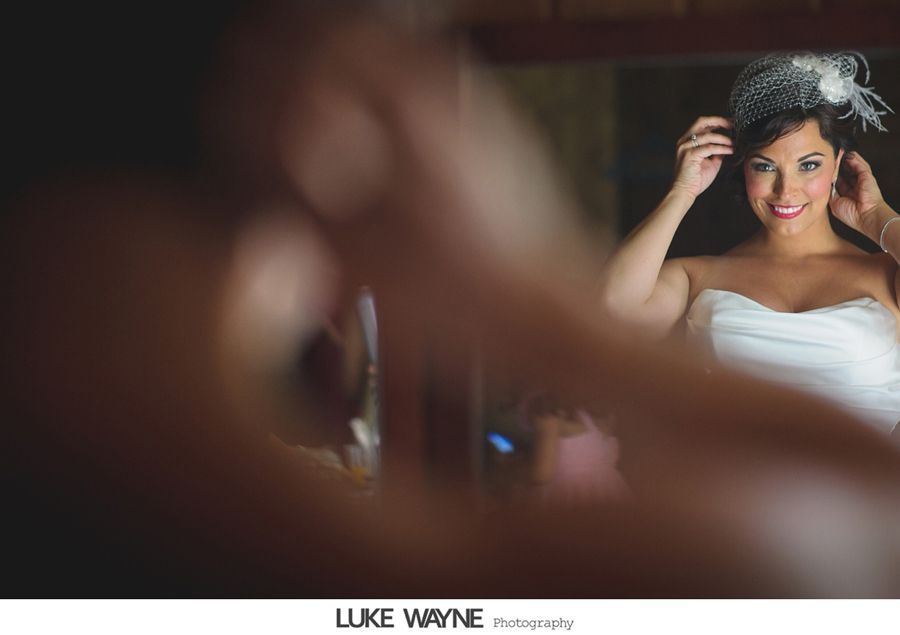 Bride adjusting headpiece in mirror, wearing strapless dress. Room with wooden accents.