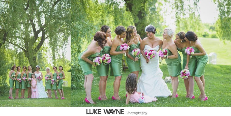 Wedding party in green dresses, pink heels, holding bouquets, with a bride in a white gown on a grassy lawn.