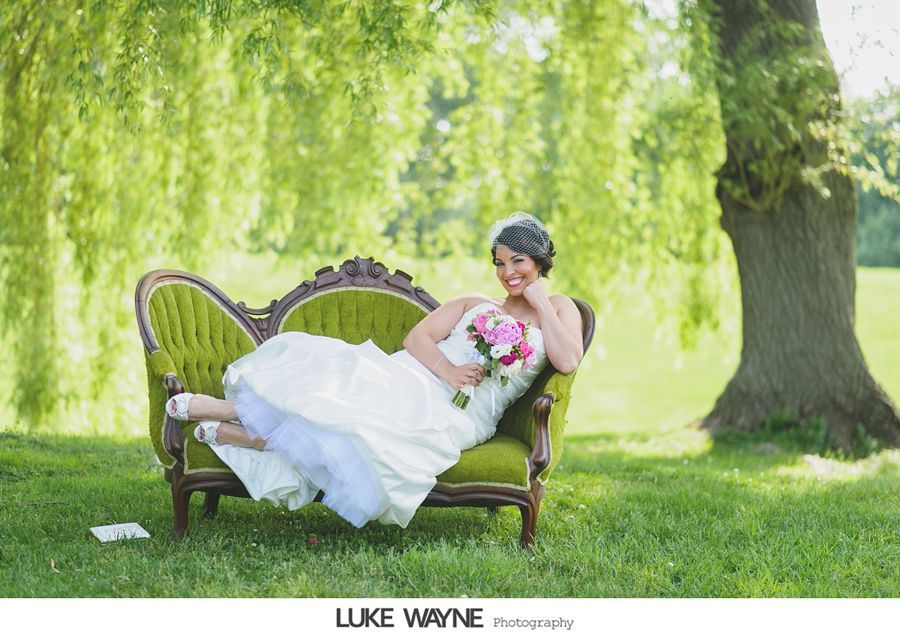 Bride in white dress reclines on green couch under a weeping willow, holding pink flowers.