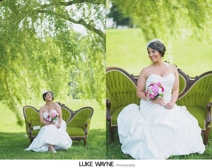 Bride in white strapless dress holding pink bouquet, sitting on a green couch in a grassy outdoor setting.