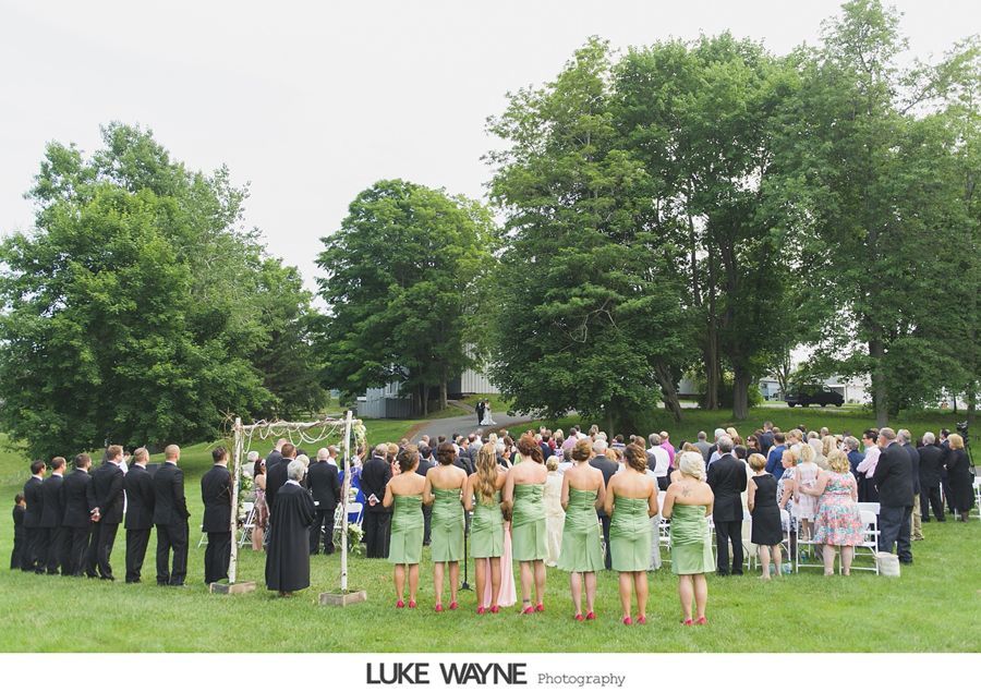 Wedding ceremony outdoors; guests stand in a grassy field. Bridesmaids in green dresses, groomsmen in black suits.