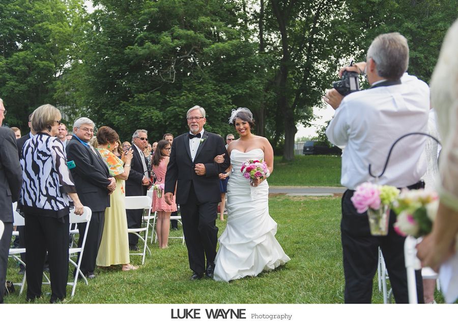 Bride and father walking down outdoor aisle; guests watch, photographer takes photo.