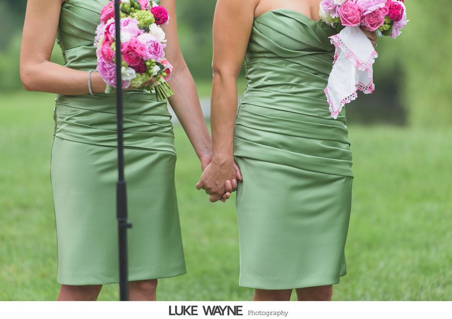 Two women in green dresses holding hands and bouquets at an outdoor event.