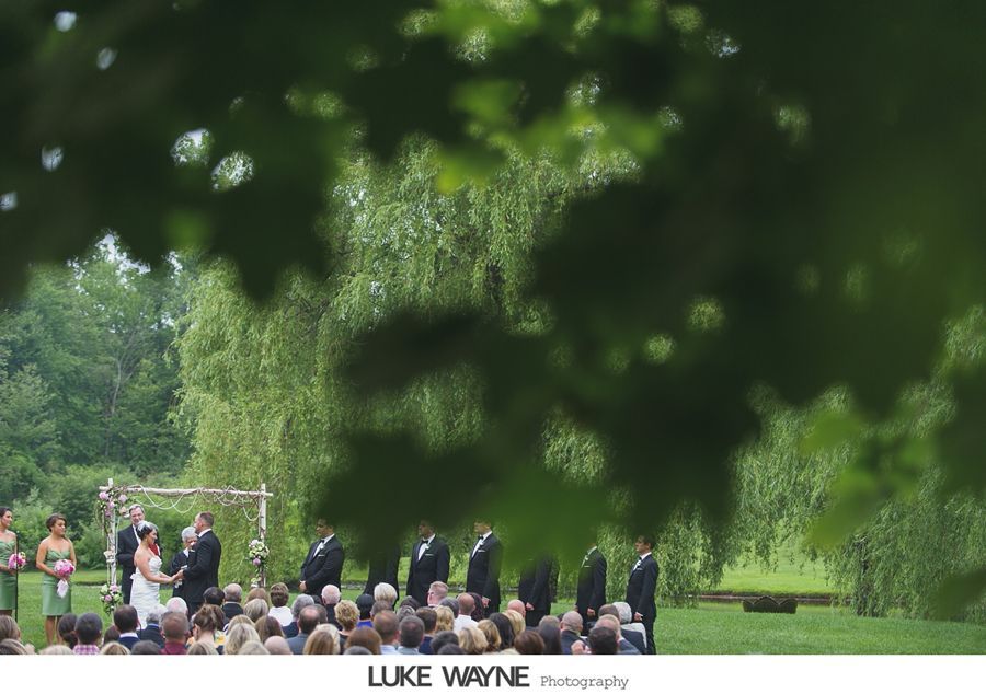 Wedding ceremony under a wooden arch, viewed through green foliage. Groom, bride, and wedding party on a grassy lawn.