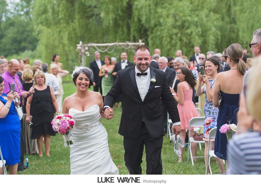 Newlyweds hold hands and smile, walking down a grassy aisle after their outdoor wedding ceremony.