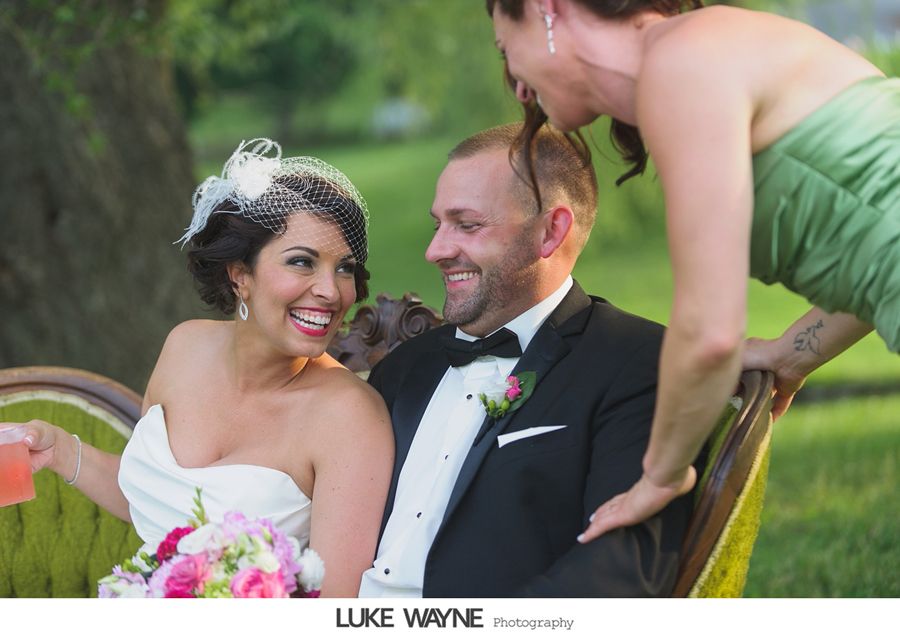 Bride and groom laugh on a vintage couch as bridesmaid playfully pulls the groom's hair.