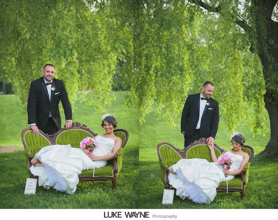 Wedding couple posing on a green vintage couch under a tree. Man in tuxedo, woman in white dress holding flowers.