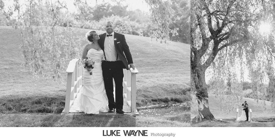 Newlyweds pose on a small bridge in a park. A large tree shades them.