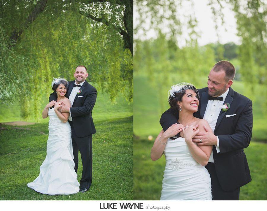 Bride and groom embrace outdoors, under a tree. She smiles, he looks at her, both in formal attire.