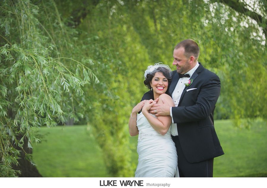 Newlyweds embrace under a weeping willow tree, smiling.