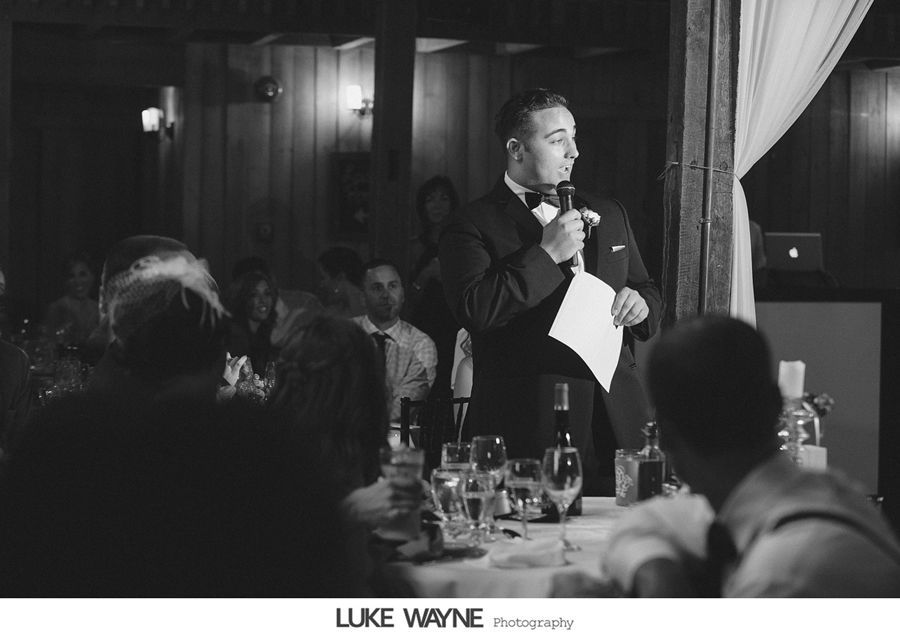 Man in tuxedo gives a speech at a wedding reception, holding paper, in a wood-paneled room.