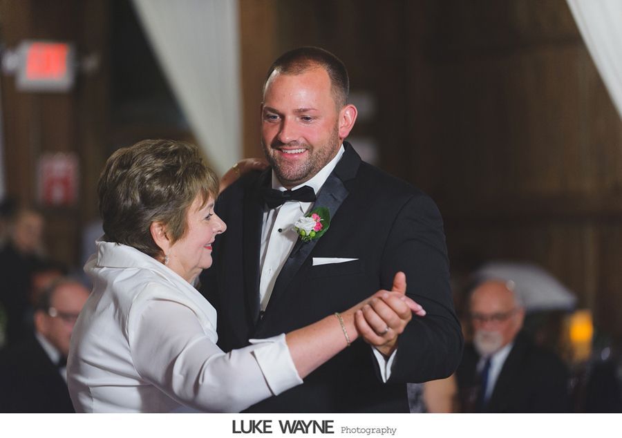 Groom in a tuxedo dances with a woman in a white jacket, indoors at a reception.