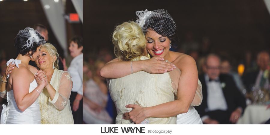 Bride hugs mother, both smiling. Wedding reception setting, warm lighting.