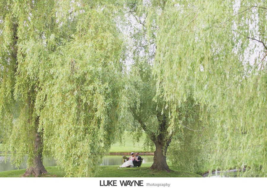 Couple sitting under weeping willow trees near a pond, dressed for a wedding.