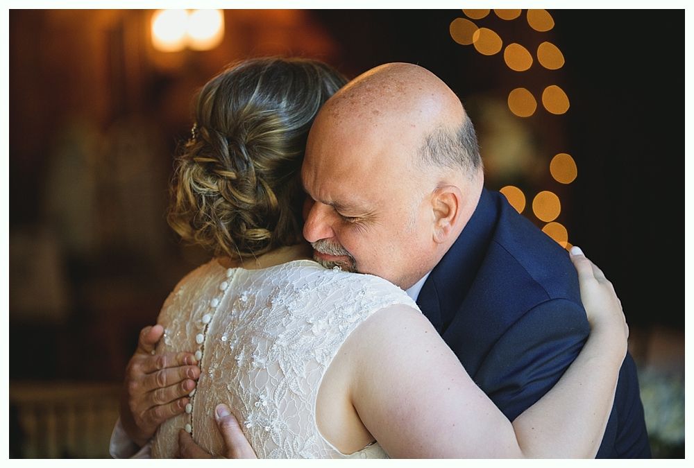 Man in a suit hugs a person in a white lace dress; they embrace. Blurry lights in background.