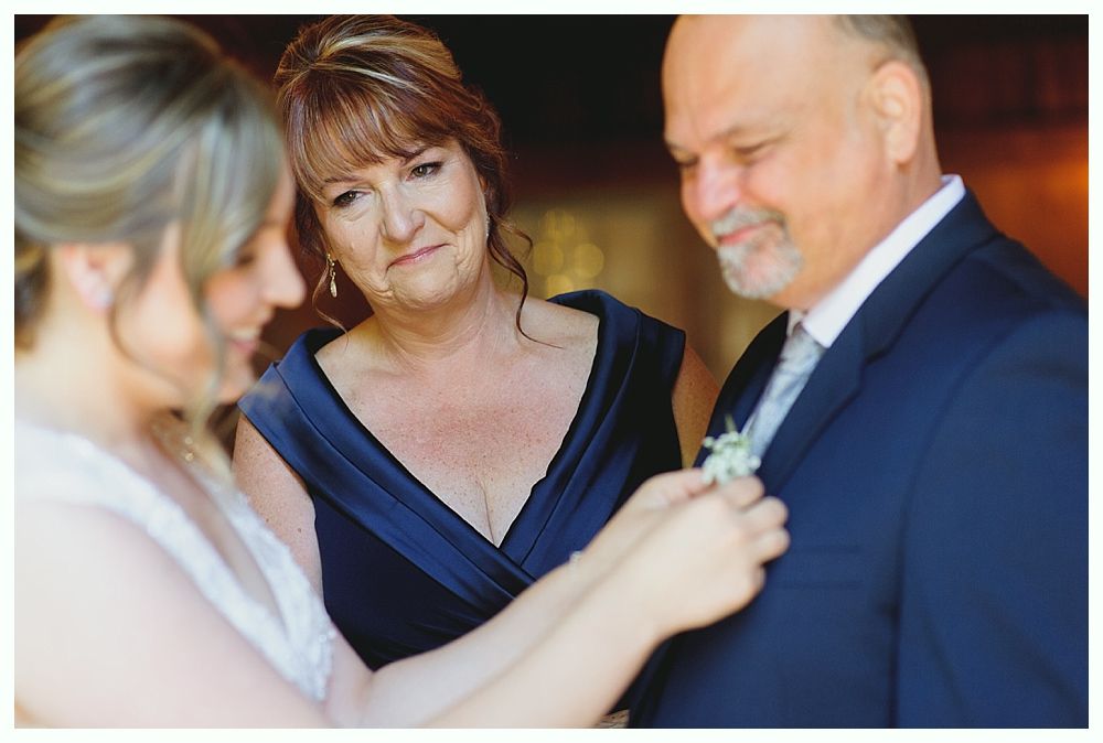 Bride pinning boutonniere on father's suit while mother watches, all in formal attire.