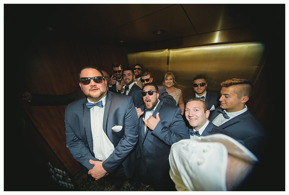 Group of men in tuxedos, sunglasses, in an elevator, posing for a photo.