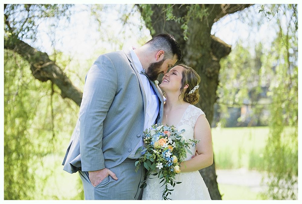 Bride and groom kissing outdoors; she holds bouquet, he wears gray suit, tree in background.