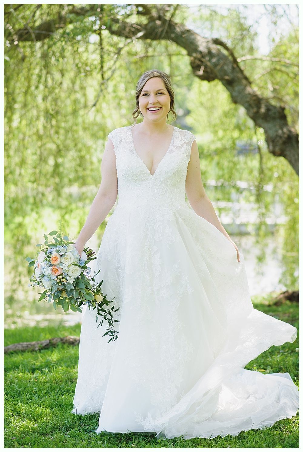 Bride in a white wedding dress holds a bouquet, smiles outdoors near a tree and water.
