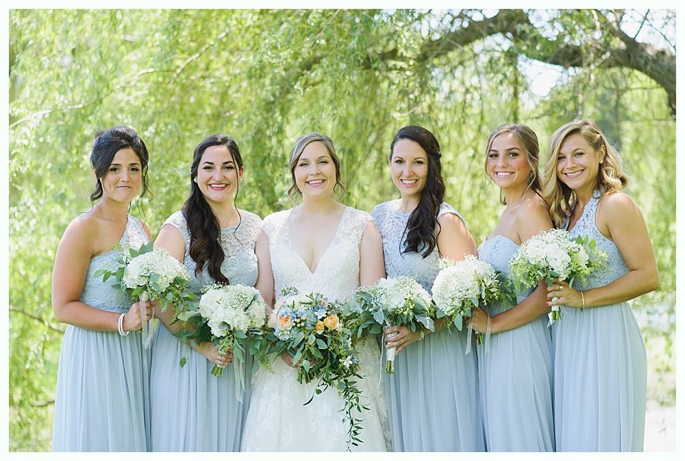 Bride and bridesmaids in light blue dresses holding bouquets, smiling outdoors.