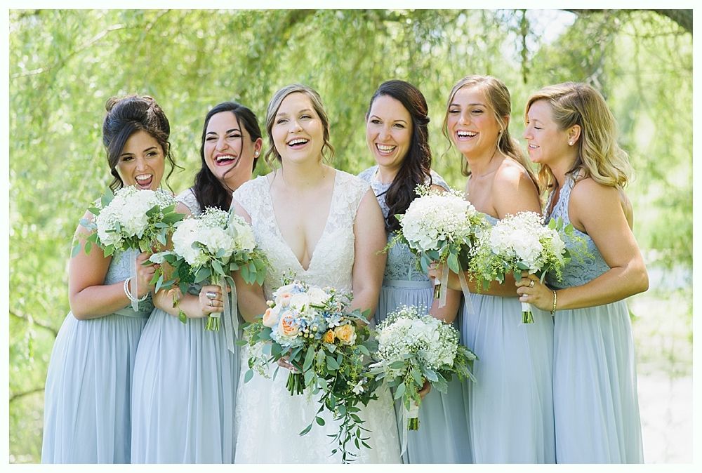 Bride and bridesmaids laugh, holding bouquets. They wear blue dresses and stand outdoors.
