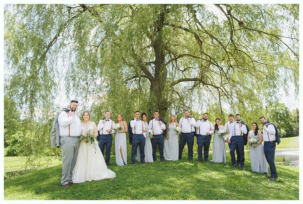 Wedding party posing under a large tree. Bride and groom centered. Groomsmen in dark pants, white shirts and suspenders. Bridesmaids in blue dresses.