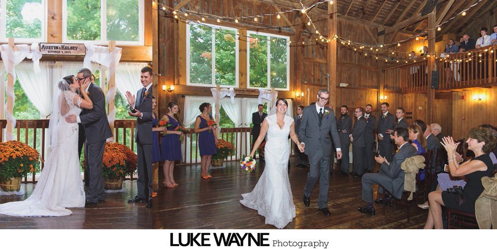 Couple kissing under a large, autumn-colored tree on a sunny, grassy hill.