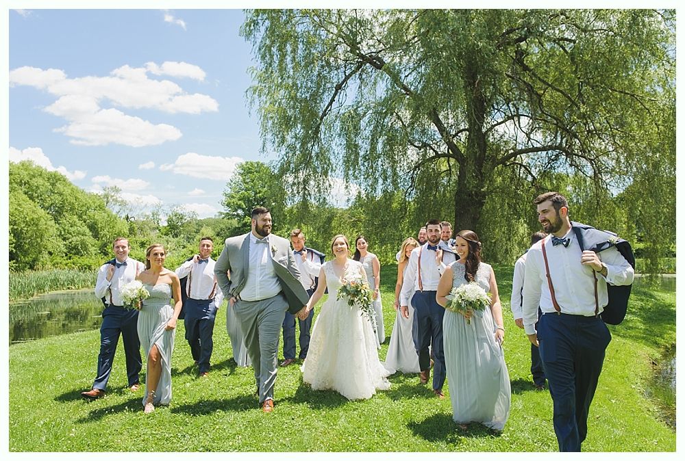 Wedding party walking on grass near a pond, under a willow tree. Blue sky with clouds.