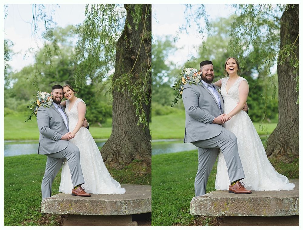 Wedding couple posing outdoors; man playfully lifts leg, woman laughs, near a tree, pond in background.