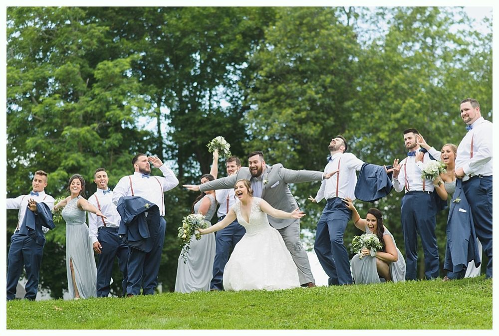 Wedding party on a grassy hill; bride and groom in center, arms outstretched. Others cheer, wearing gray and blue.